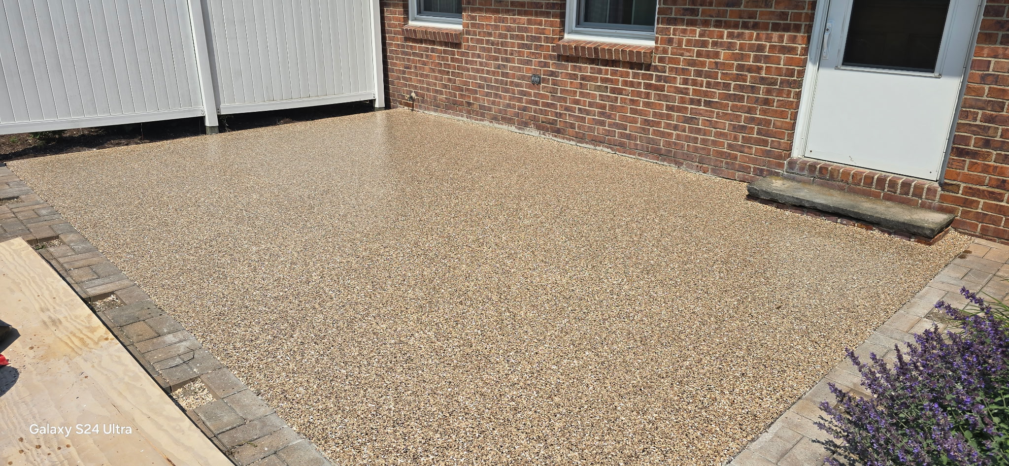 Tan decorative flake patio flooring beside a brick home and white privacy fence